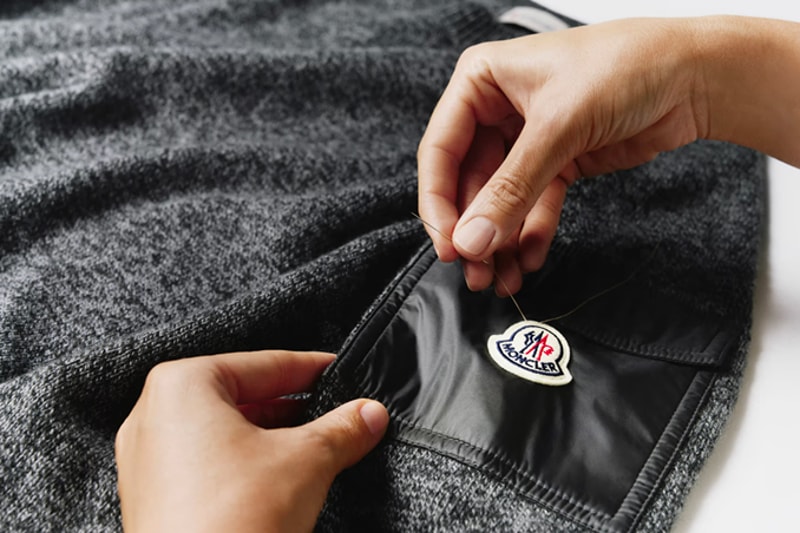 Hands examining the Moncler logo patch on a grey knit garment as part of the product aftercare service.