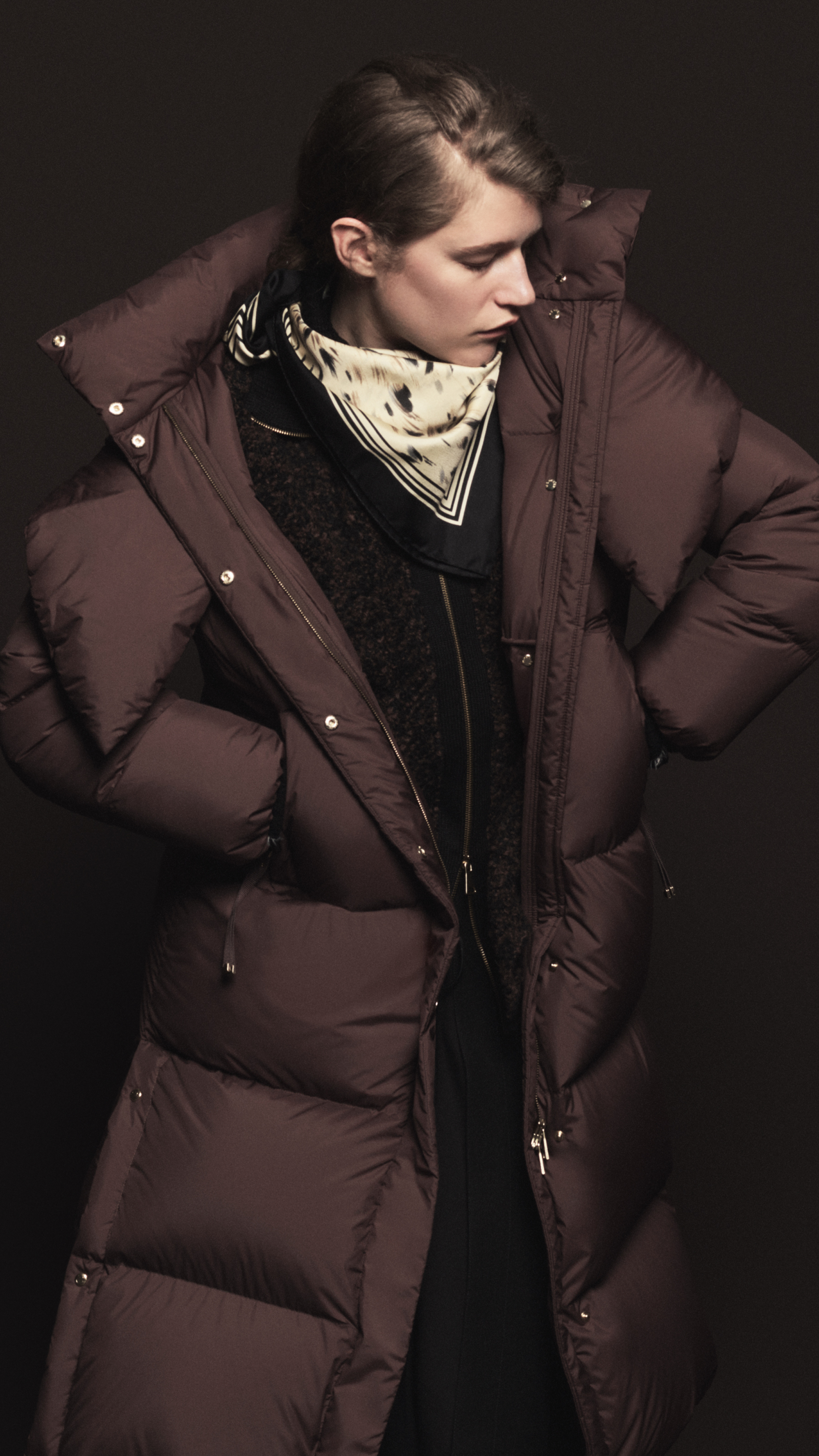 Moncler Fall/Winter 2026 for men and women. Oversized burgundy down jacket with hood, printed cream scarf and black zip knit.
